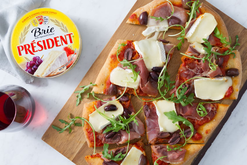 A photo by Morgan Ione for Bon Appétit Magazine featuring a circular, yellow package of Président brie next to a wooden board with a flatbread on it. The flatbread has tomato sauce, kalamata olives, thinly sliced meats, arugula, and slices of brie. There is a a stemless glass of red wine partially out of frame.